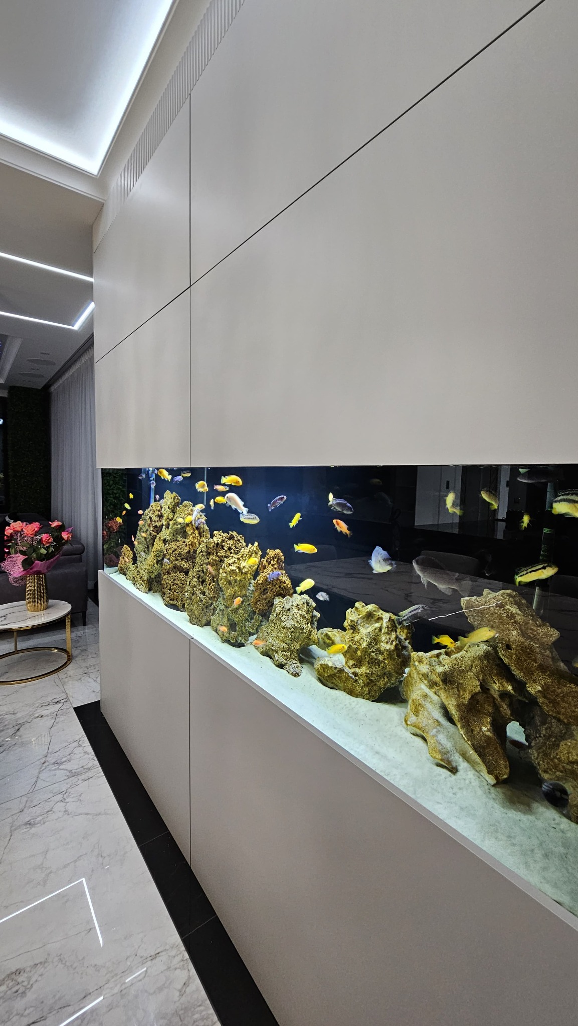 Nowoczesny salon z wbudowanym akwarium z rybami, skałami i piaskiem, otoczony białymi panelami ściennymi i marmurową podłogą.