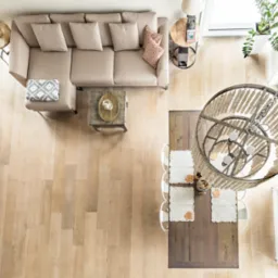 Jasna podłoga z drewna z widokiem z góry na salon: beżowa sofa, drewniany stół z dekoracjami, schody betonowe. Naturalne światło podkreśla teksturę drewna.