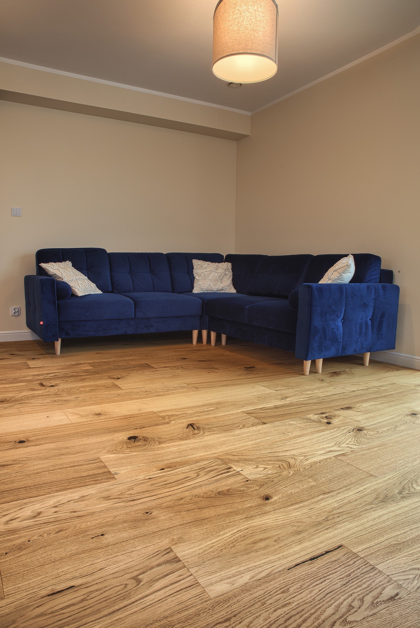 Jasna, drewniana podłoga ułożona w jodełkę francuską, uzupełniona o granatową, welurową sofę narożną i lampę sufitową. Wnętrze w neutralnej kolorystyce.