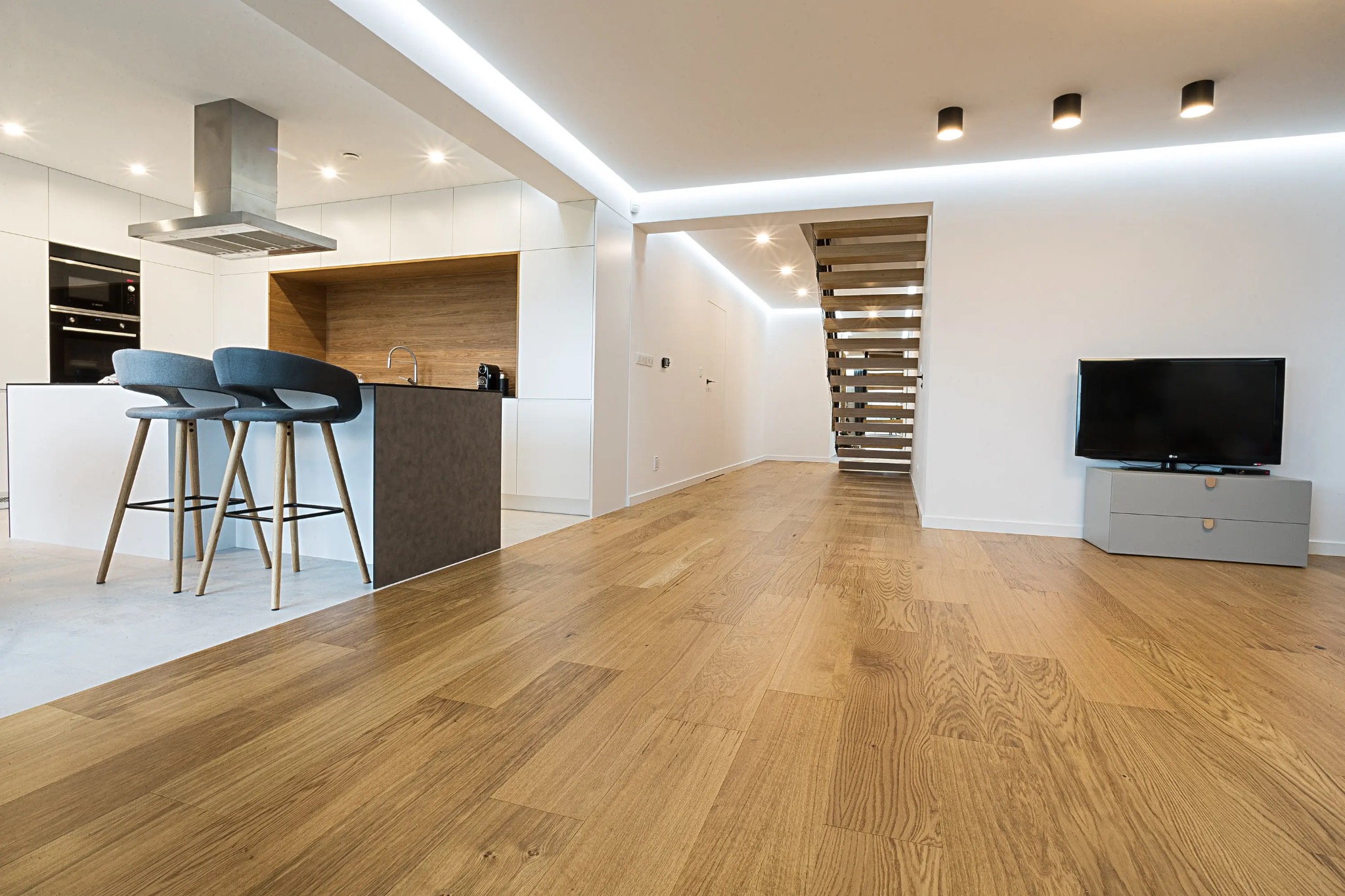Jasne drewno na podłodze w nowoczesnym wnętrzu. Widok na otwartą kuchnię z szarymi stołkami barowymi i schody. Minimalistyczny design, ciepłe oświetlenie.