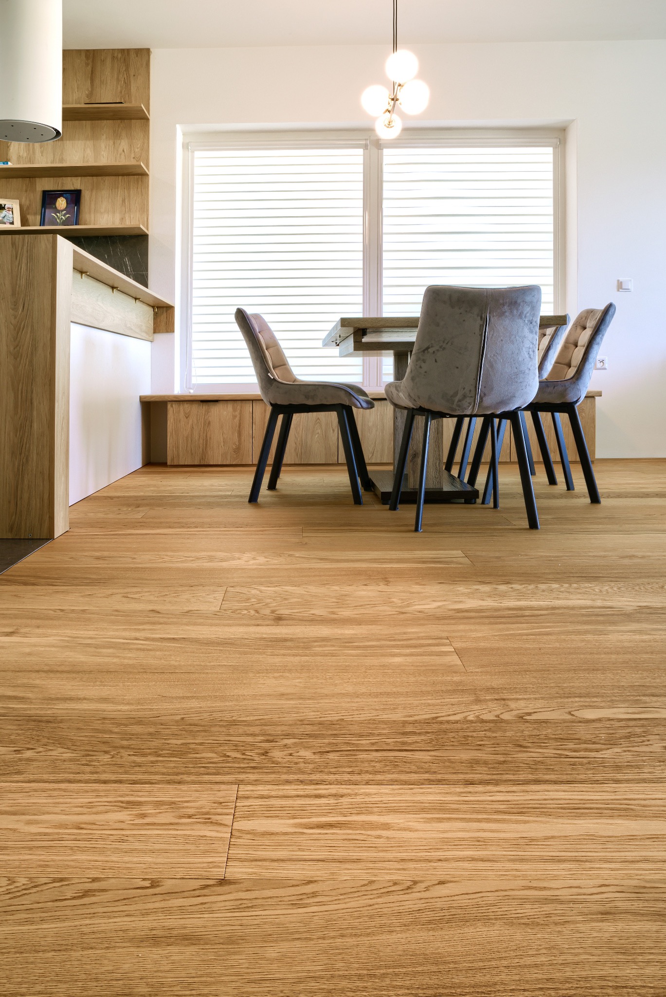 Jasna dębowa posadzka w nowoczesnej jadalni. Widoczne krzesła z szarej tkaniny i drewniany stół pod oknem z żaluzjami. Minimalistyczne wnętrze z drewnianymi elementami.