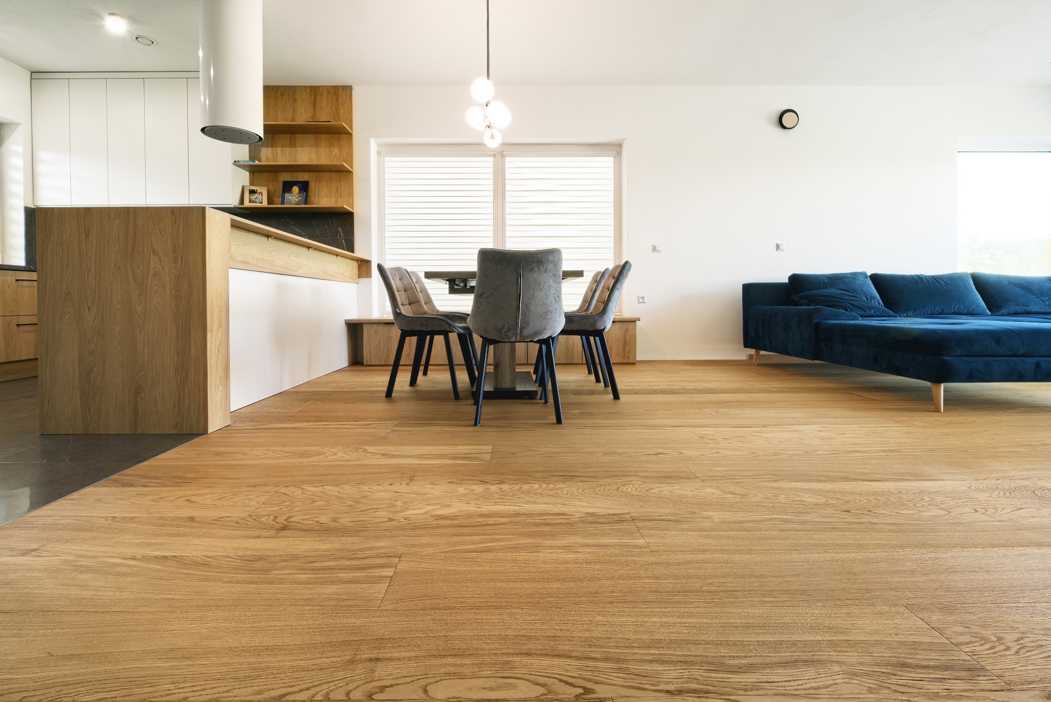 Jasna, drewniana podłoga w nowoczesnym salonie z kuchnią; widoczny fragment niebieskiej sofy, stół z krzesłami i elementy zabudowy kuchennej.
