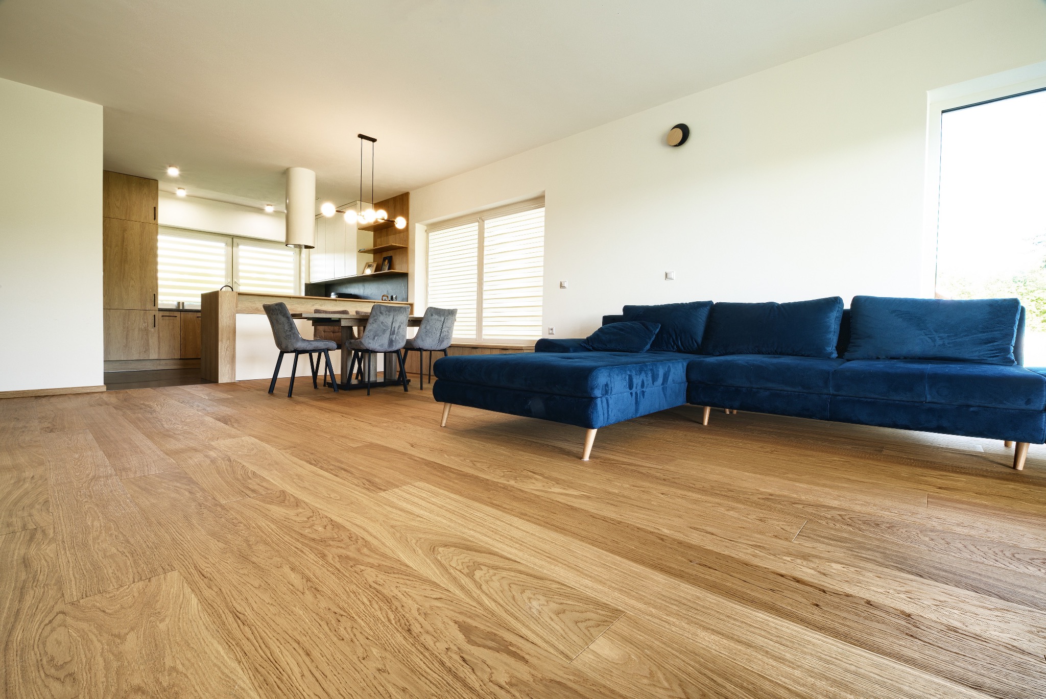 Jasne deski w salonie z aneksem kuchennym. W tle stół z krzesłami i niebieski narożnik. Widok na ułożoną podłogę z perspektywy podłogi.