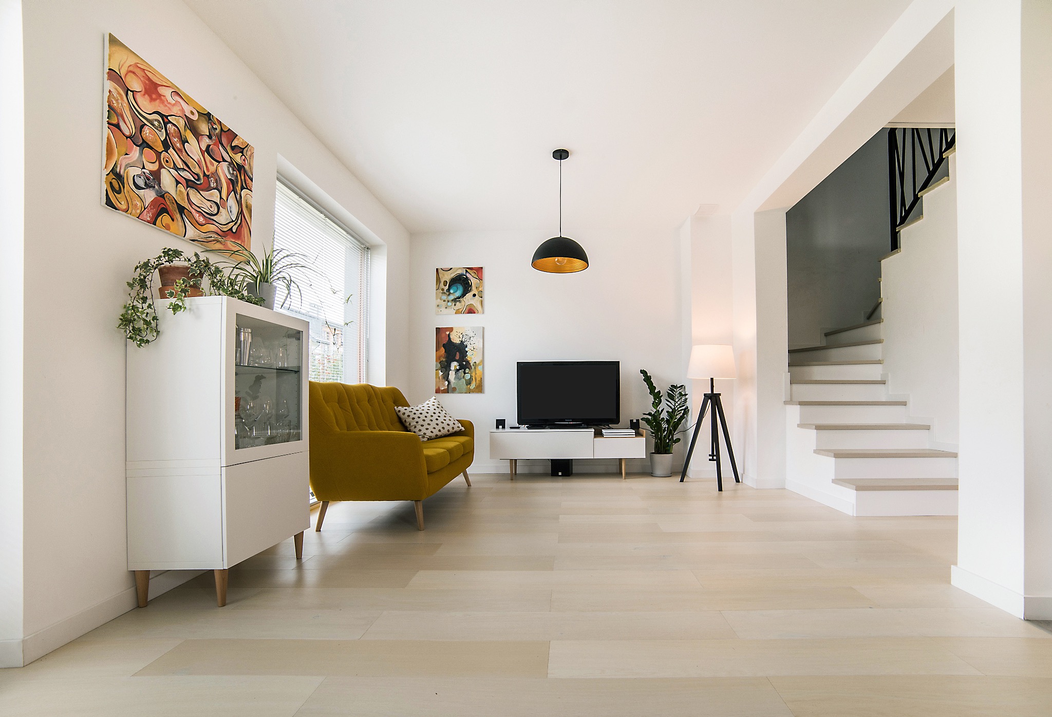 Jasne, drewniane deski podłogowe w przestronnym salonie z żółtą sofą, białymi meblami i schodami. Nowoczesne oświetlenie i dekoracje.