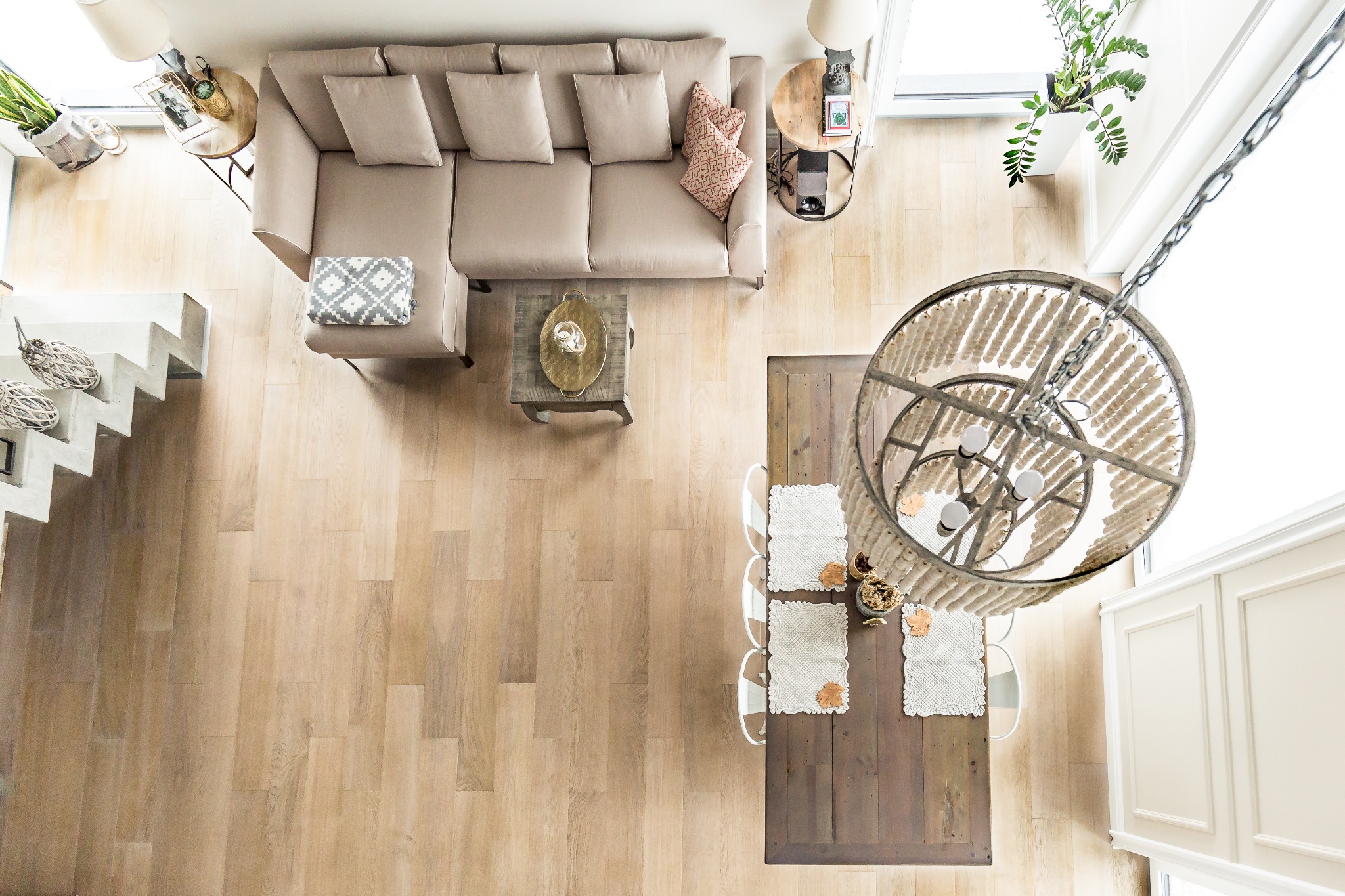 Jasna podłoga z drewna z widokiem z góry na salon: beżowa sofa, drewniany stół z dekoracjami, schody betonowe. Naturalne światło podkreśla teksturę drewna.