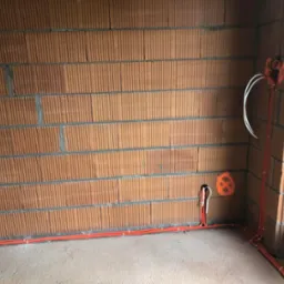 Pomarańczowe rury instalacyjne ułożone wzdłuż ceglanej ściany w surowym stanie, z wystającymi białymi kablami i pomarańczowym oznaczeniem na ścianie, widoczne okno z naklejkami.