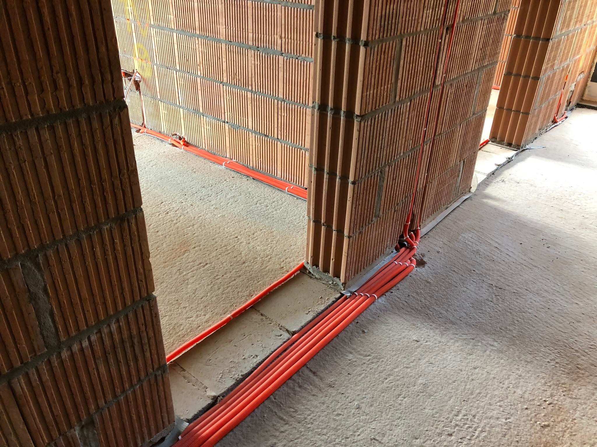 Pomarańczowe rury instalacyjne ułożone w bruzdach w ścianie z pustaków ceramicznych, widok z perspektywy podłogi w surowym stanie.