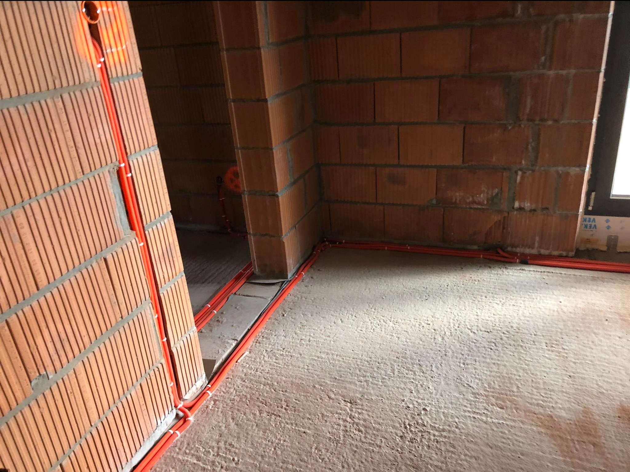 Pomieszczenie w surowym stanie z widocznymi pomarańczowymi rurami instalacji elektrycznej, ułożonymi wzdłuż ścian i podłogi, na tle ceglanych ścian i betonowej posadzki.