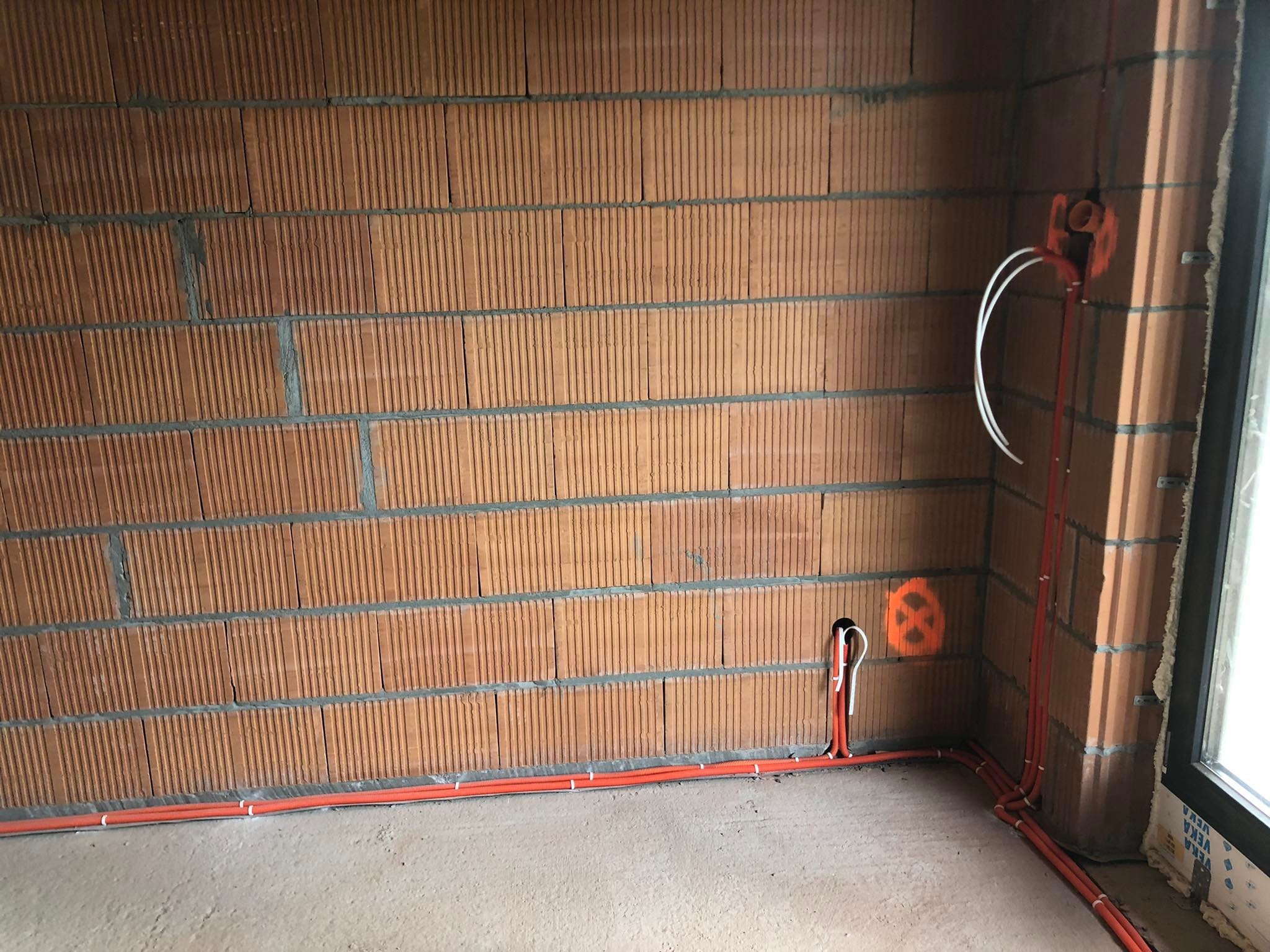 Pomarańczowe rury instalacyjne ułożone wzdłuż ceglanej ściany w surowym stanie, z wystającymi białymi kablami i pomarańczowym oznaczeniem na ścianie, widoczne okno z naklejkami.