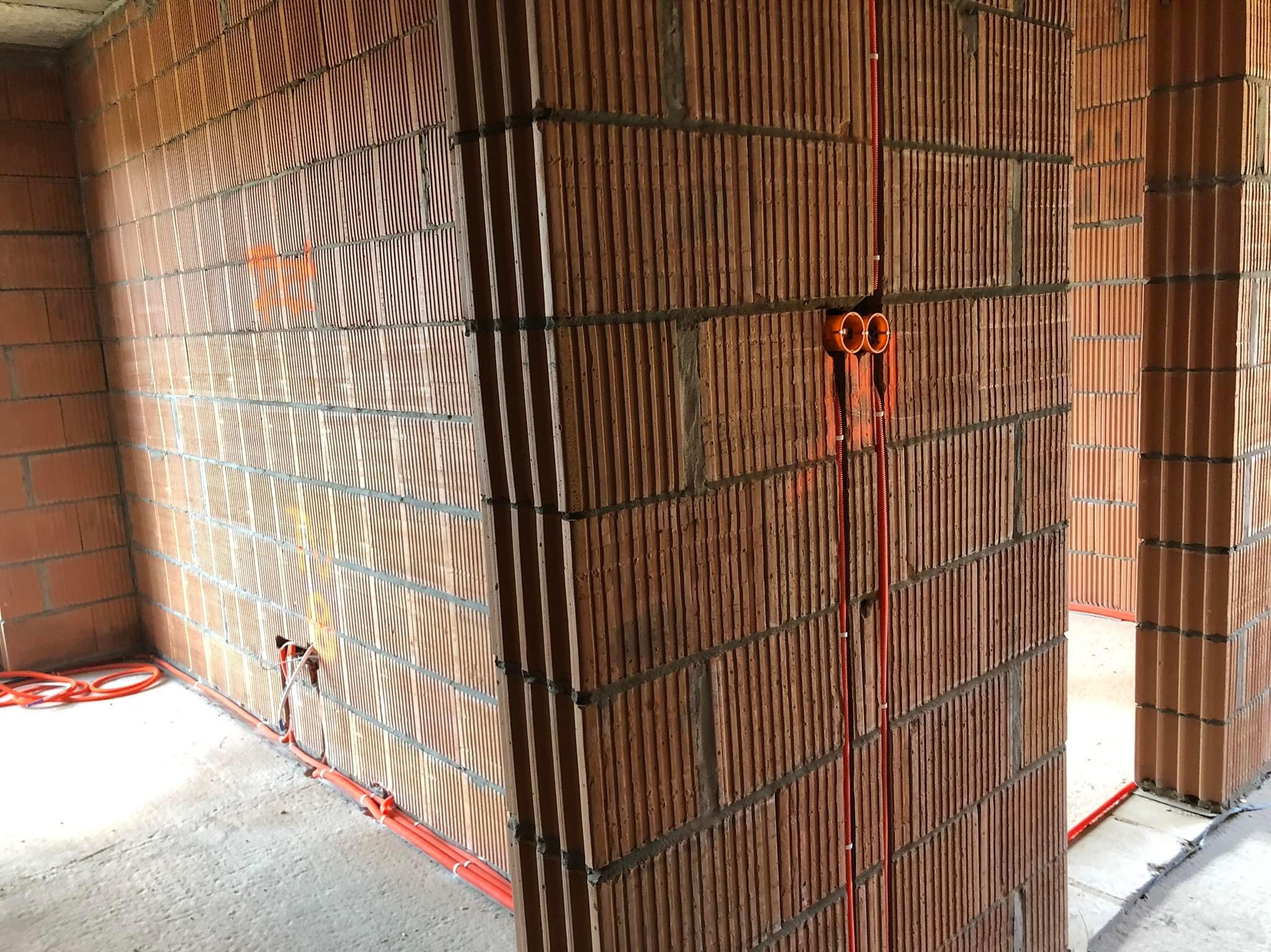 Pomarańczowe rury instalacyjne, prowadzone pionowo i poziomo po ścianie z cegieł ceramicznych dziurawki w surowym stanie, z widocznymi zaprawami i oznaczeniami na ścianie.