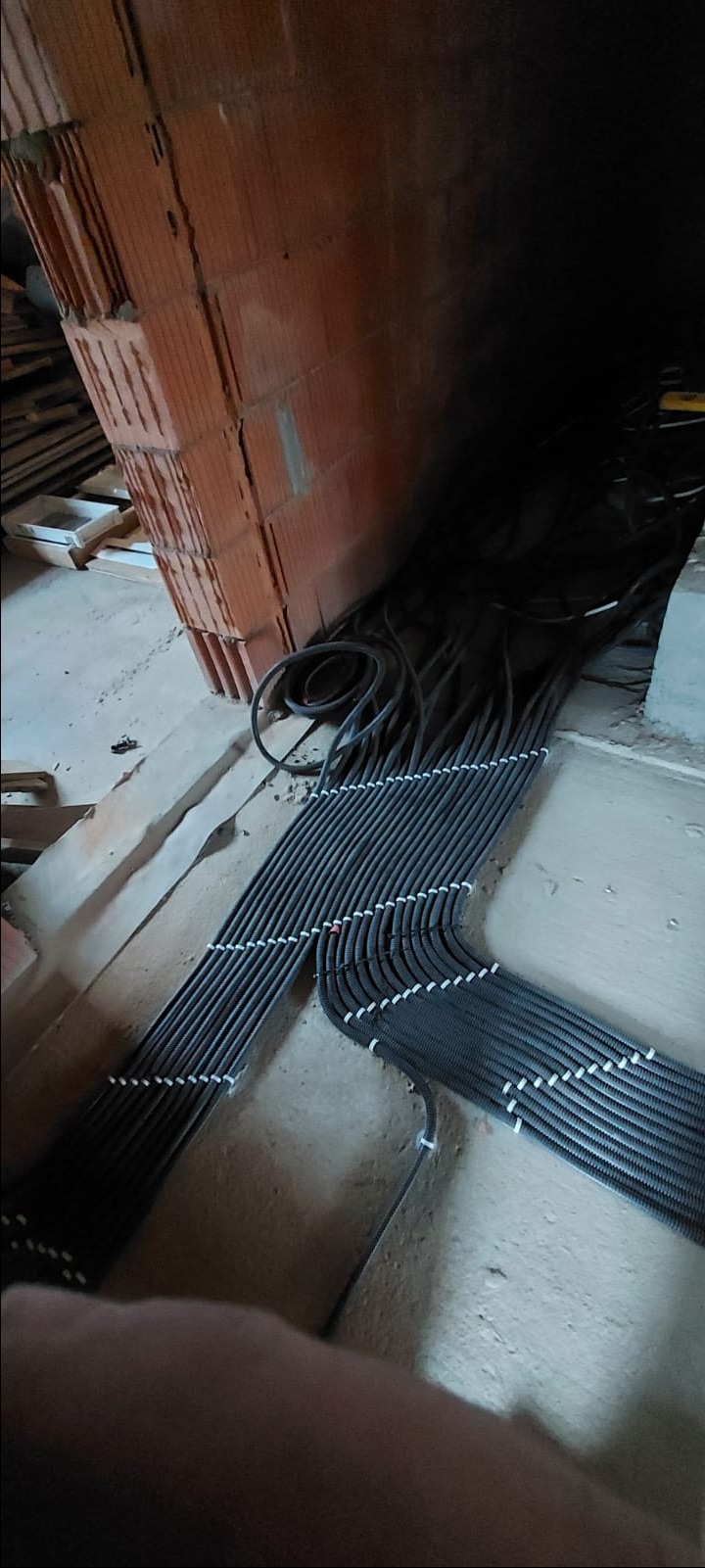 Równolegle ułożone, czarne, karbowane rury instalacyjne z białymi uchwytami mocującymi, prowadzone po betonowej podłodze w surowym stanie, w tle fragment ściany z czerwonej cegły.