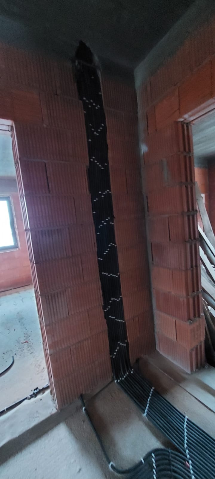 Pionowy układ kabli elektrycznych, zabezpieczonych uchwytami, w bruździe wymurowanej ściany z pustaków ceramicznych na surowym etapie budowy.