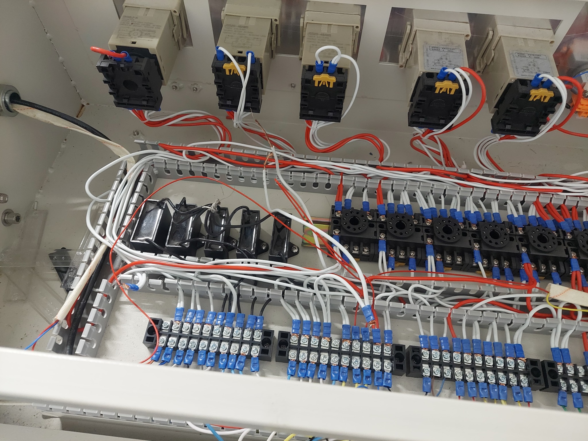 Wnętrze skrzynki elektrycznej z przekaźnikami, bezpiecznikami i okablowaniem w kolorach białym, czerwonym i niebieskim, widoczne ślady montażu i podłączeń.