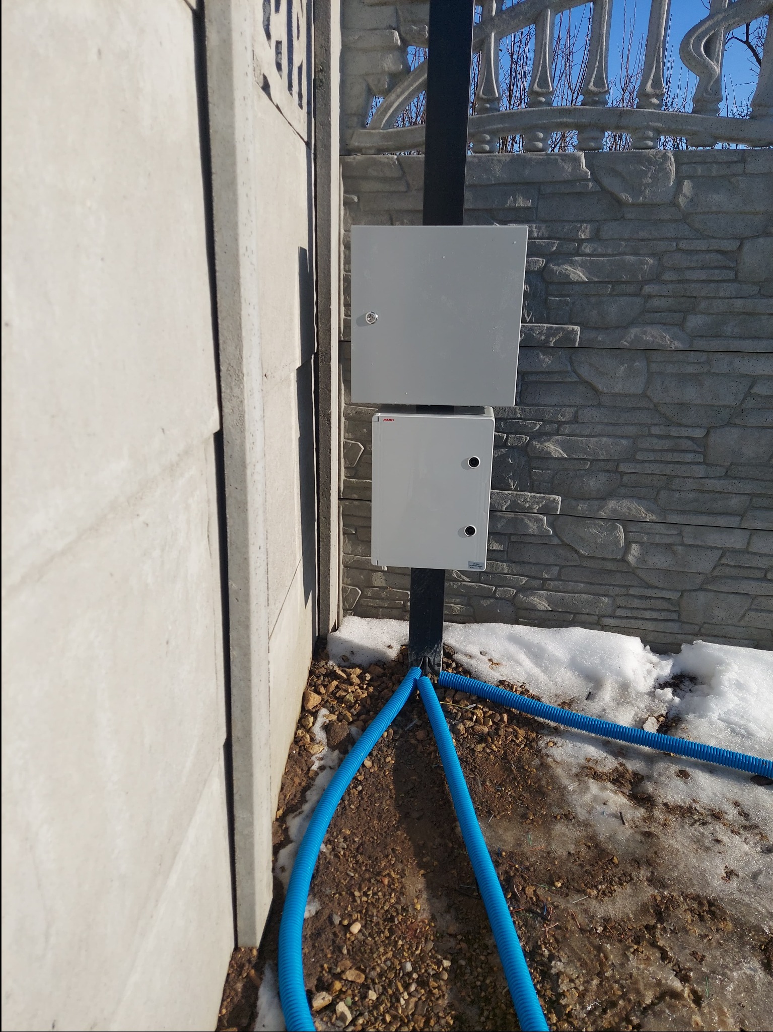 Dwie szare skrzynki elektryczne zamontowane na czarnym słupku, z niebieskimi rurami karbowanymi prowadzącymi do ziemi, na tle betonowego ogrodzenia z dekoracyjnym wzorem i resztkami śniegu.