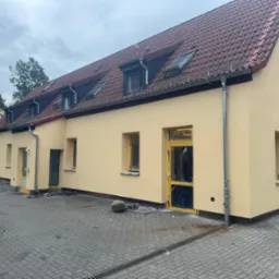 Dwukondygnacyjny budynek z nową, jasnobeżową elewacją, czerwonym dachem z oknami dachowymi i szarą kostką brukową na podjeździe. Okna i drzwi zabezpieczone folią malarską.