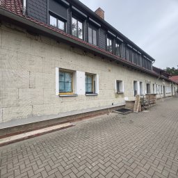 G-BAUMONTSYSTEMS SPÓŁKA Z OGRANICZONĄ ODPOWIEDZIALNOŚCIĄ - Ocieplanie elewacji budynku wielorodzinnego z widoczną warstwą izolacyjną z wełny mineralnej i przygotowanymi otworami okiennymi, widok z poziomu brukowanego chodnika.