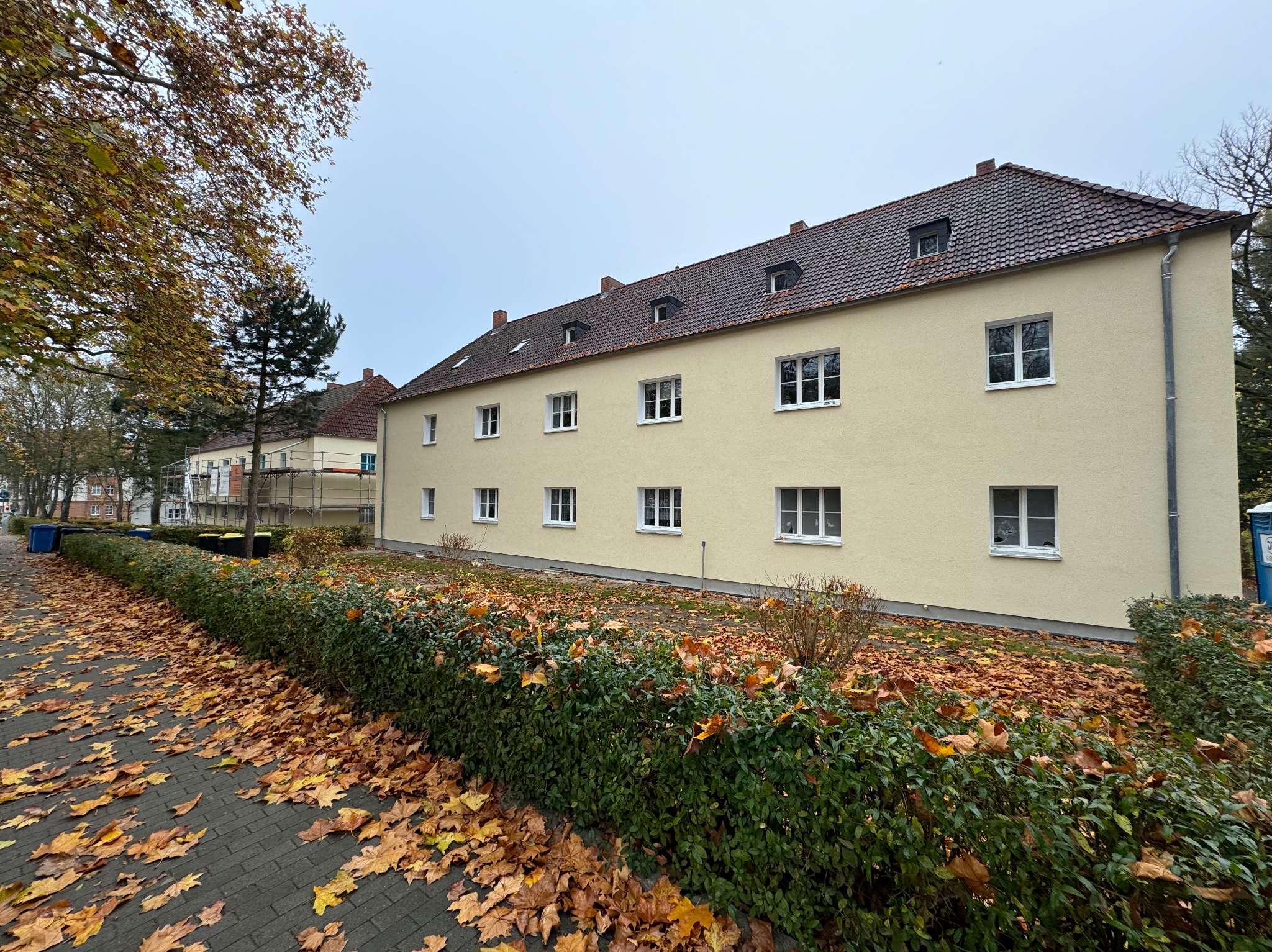 Żółty budynek wielorodzinny z brązowym dachem i białymi oknami, widoczny z chodnika pokrytego opadłymi liśćmi, z krzewami wzdłuż budynku i fragmentem drugiego budynku w tle.