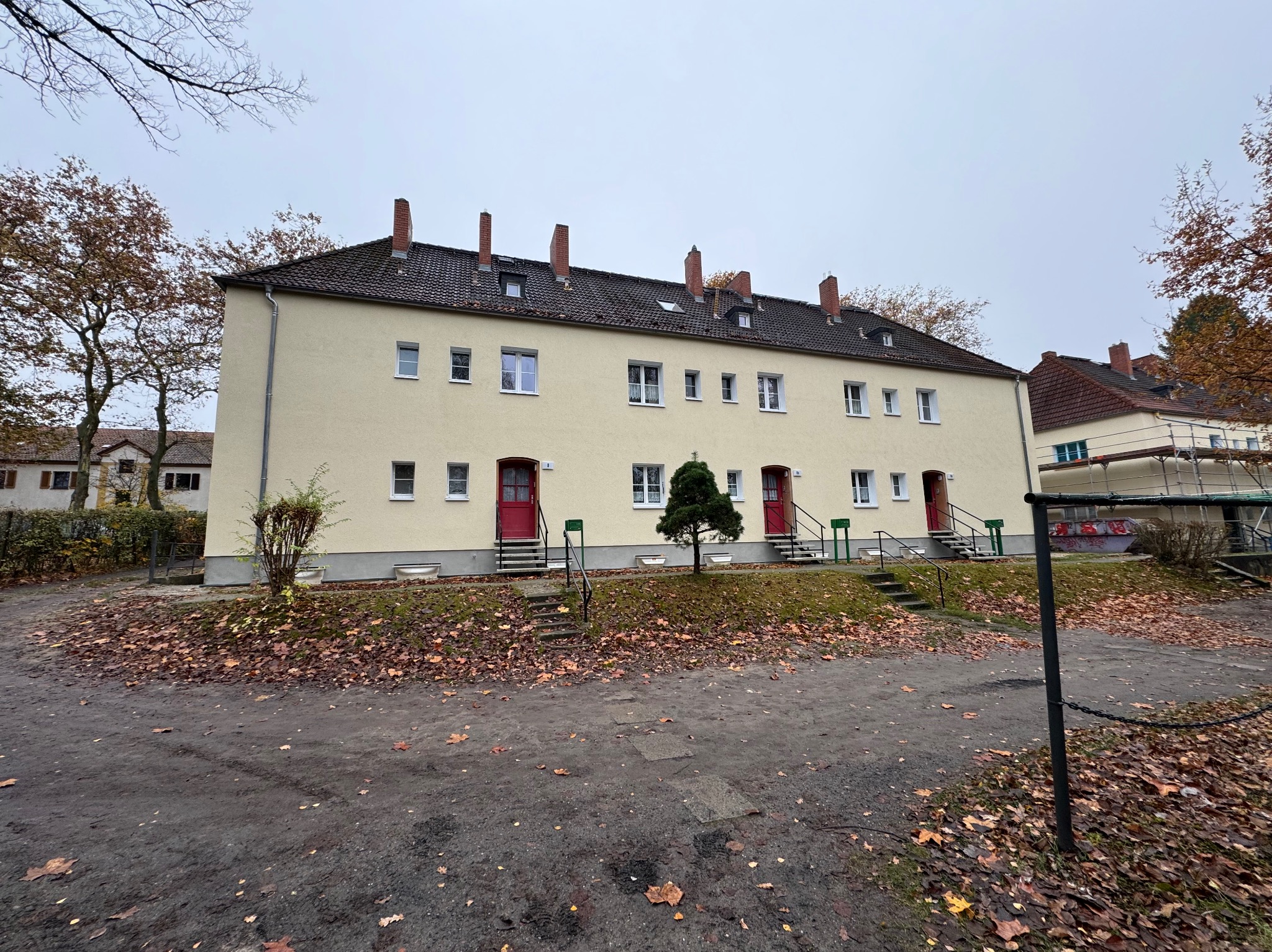 Żółta elewacja szeregowca z czerwonymi drzwiami wejściowymi, widok z poziomu gruntu, jesienne liście na trawniku.