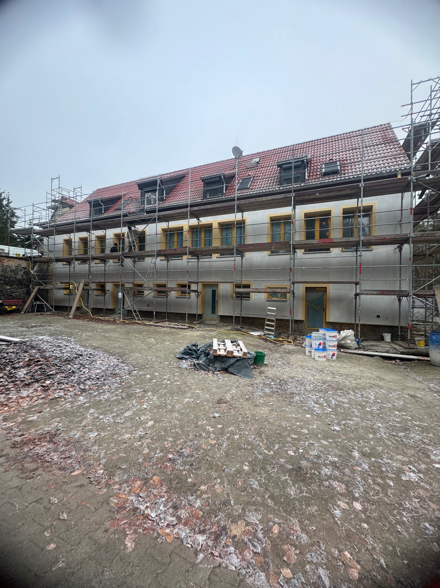 Dwupiętrowy budynek w trakcie prac elewacyjnych z widocznymi rusztowaniami, częściowo otynkowaną fasadą, stertą liści na ziemi i wiadrami z farbą u podstawy.
