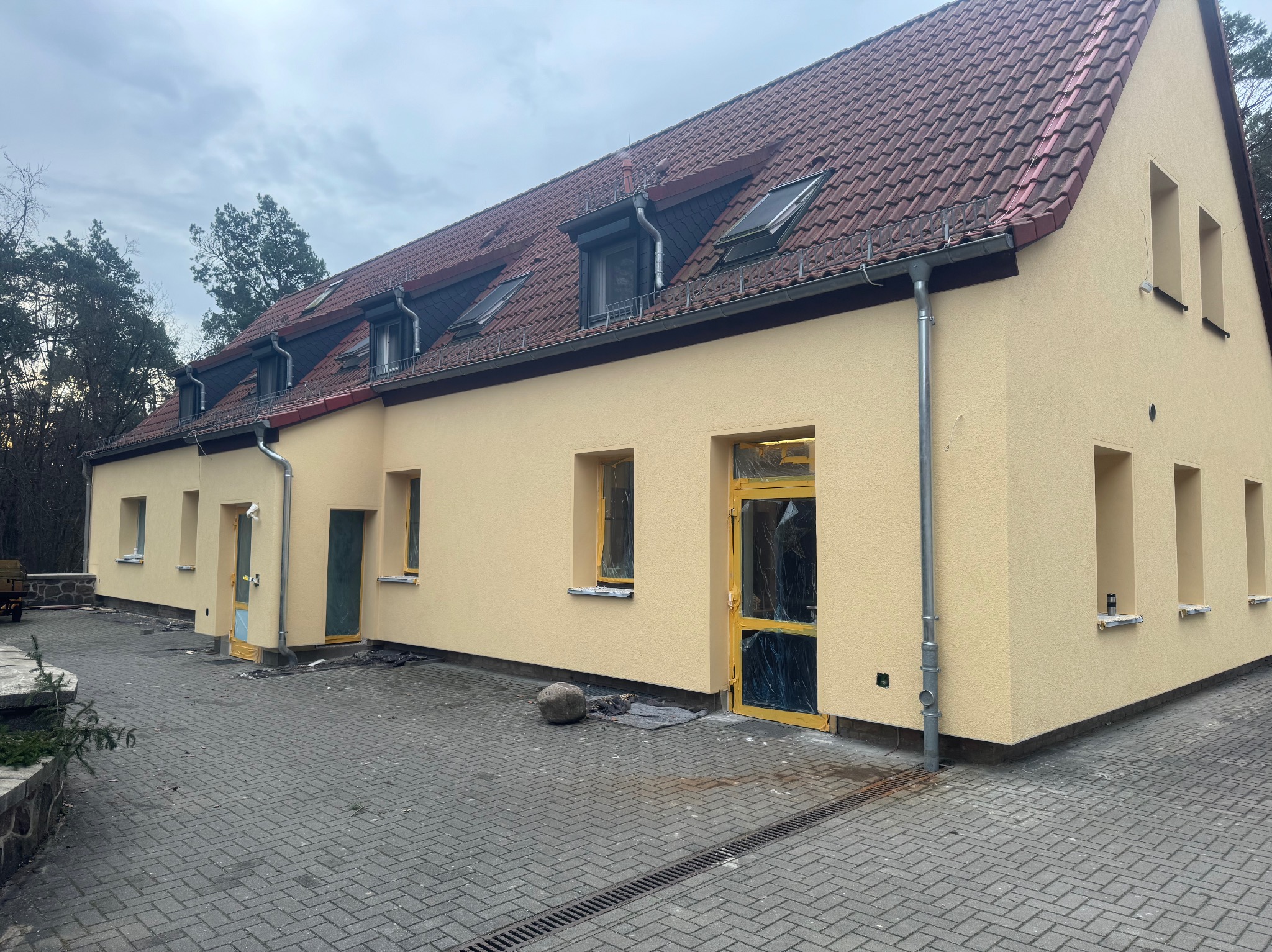 Dwukondygnacyjny budynek z nową, jasnobeżową elewacją, czerwonym dachem z oknami dachowymi i szarą kostką brukową na podjeździe. Okna i drzwi zabezpieczone folią malarską.