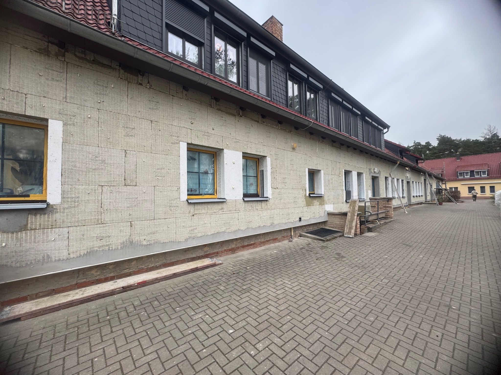 Ocieplanie elewacji budynku wielorodzinnego z widoczną warstwą izolacyjną z wełny mineralnej i przygotowanymi otworami okiennymi, widok z poziomu brukowanego chodnika.