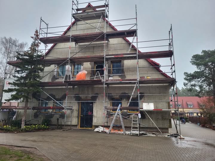 Dom jednorodzinny w trakcie prac elewacyjnych, widoczne rusztowania, zabezpieczone okna i drzwi, ślady po naprawach na elewacji, drabina oparta o ścianę, żółte kwiaty przed budynkiem.