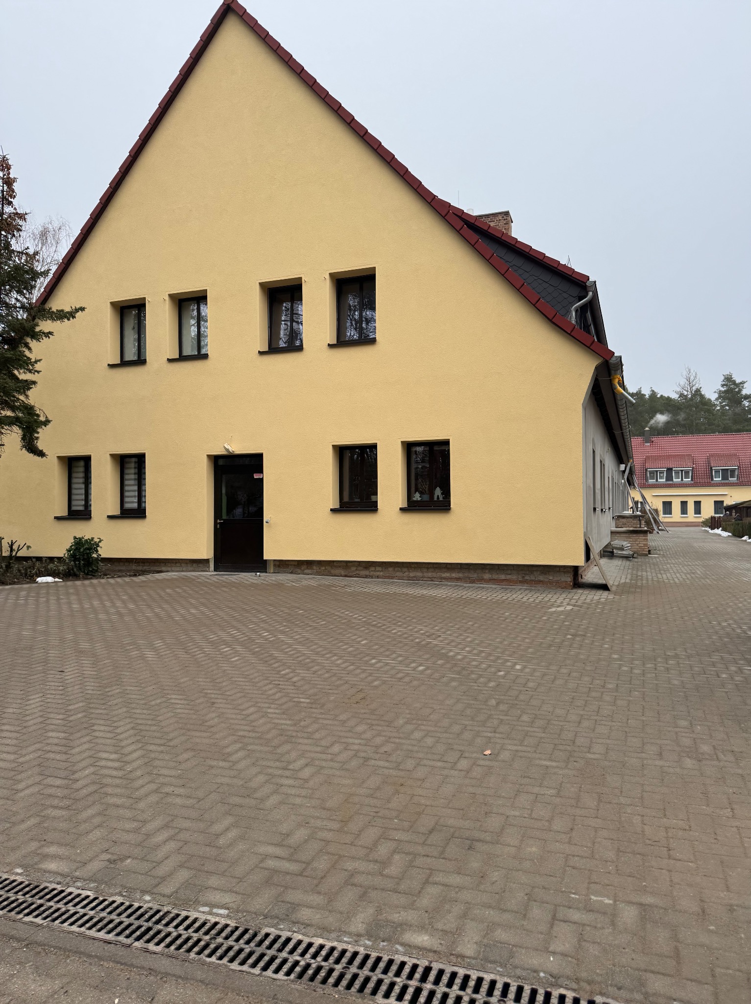 Żółta elewacja dwukondygnacyjnego budynku z czerwonym dachem, widok z brukowanego podjazdu w pochmurny dzień.