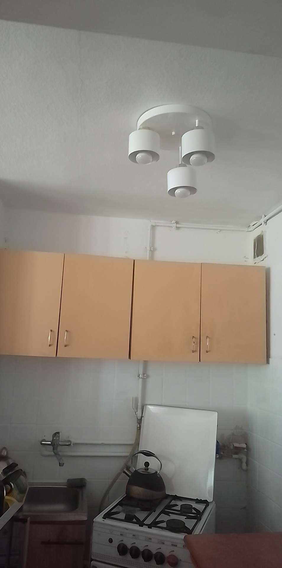 Kuchnia z beżowymi szafkami wiszącymi, białą kuchenką gazową z czajnikiem i okrągłą, trzypunktową lampą sufitową w stylu skandynawskim.