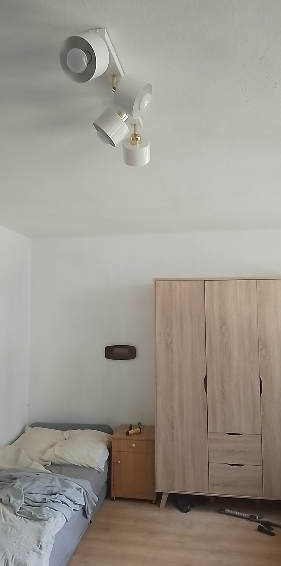 Biała lampa sufitowa z trzema reflektorami o regulowanym kącie świecenia, oświetlająca sypialnię z łóżkiem, szafką nocną i dużą szafą z jasnego drewna.