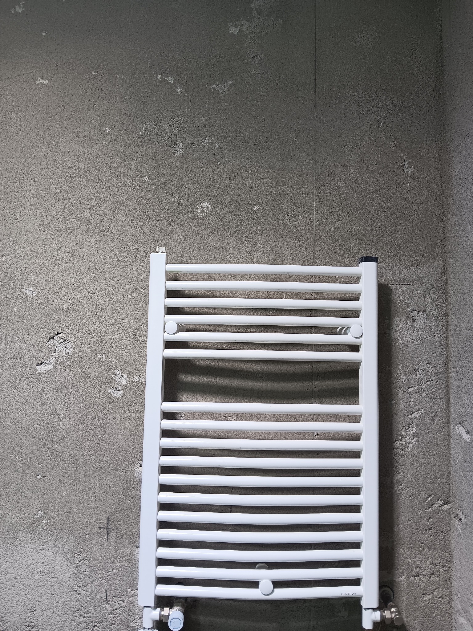Biały, drabinkowy grzejnik łazienkowy zamontowany na surowej, szarej ścianie z widocznymi śladami po tynku.