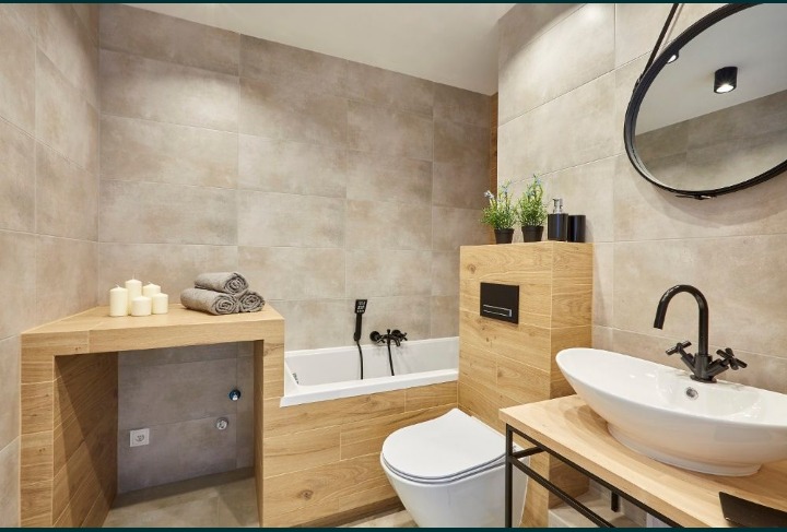 Nowoczesna łazienka z beżowymi płytkami imitującymi drewno, owalną umywalką na metalowym stelażu, wanną z czarną armaturą i okrągłym lustrem z czarną ramą.