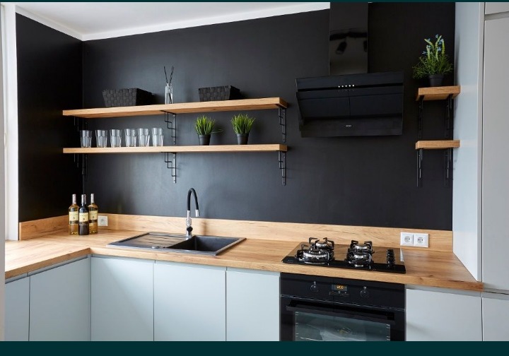 Nowoczesna kuchnia z czarną ścianą, drewnianymi półkami i jasnymi szafkami, wyposażona w czarny zlew, płytę grzewczą i piekarnik.