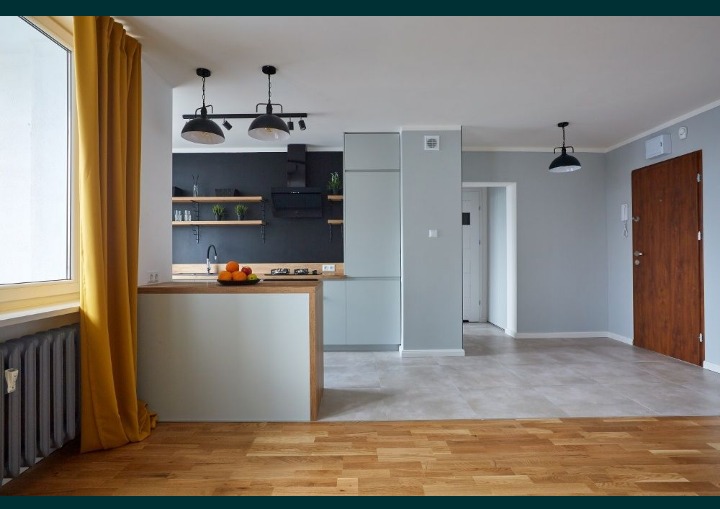 Przestronne wnętrze mieszkania po remoncie: widok na kuchnię z wyspą, drewniane podłogi przechodzące w szare płytki, żółte zasłony przy oknie i czarna ściana z półkami w tle.