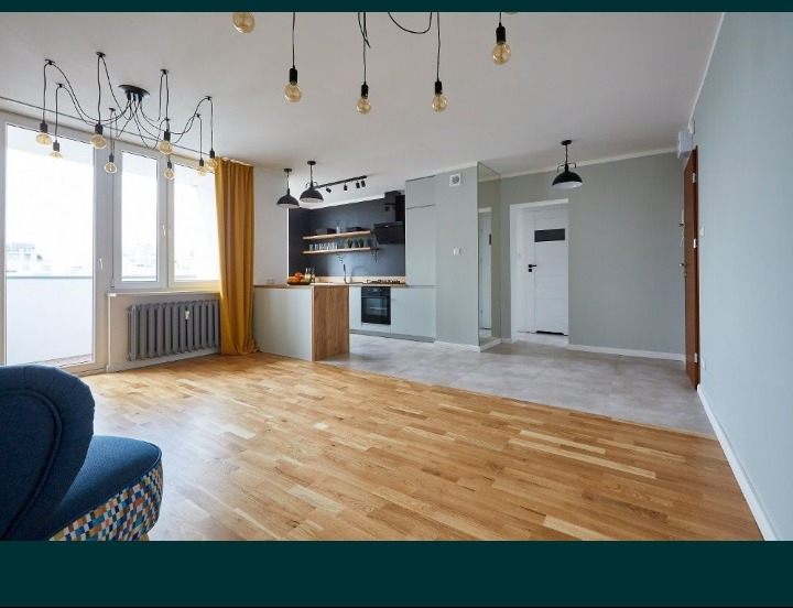 Jasne wnętrze mieszkania po remoncie: drewniana podłoga, szare ściany, otwarta kuchnia z czarną ścianą i drewnianymi półkami, widoczny fragment niebieskiego fotela z wzorzystą tapicerką...