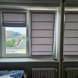 Dwa okna z szarymi tekstylnymi osłonami, jedno częściowo podniesione, widok na miasto z pociągami w tle, fragment grzejnika i szafy z lustrem w pomieszczeniu.