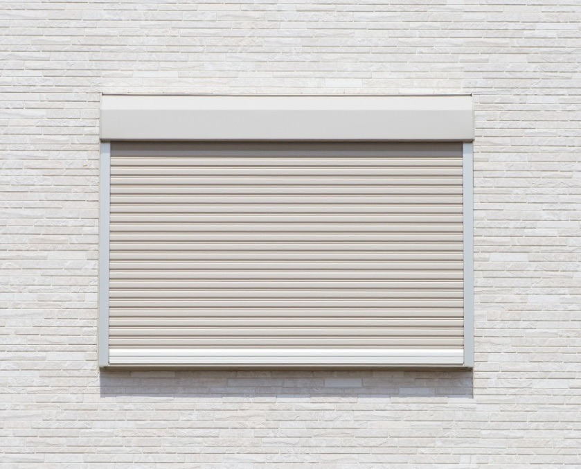 Zamknięta, beżowa osłona okienna na jasnej, ceglanej ścianie budynku, widok z zewnątrz.
