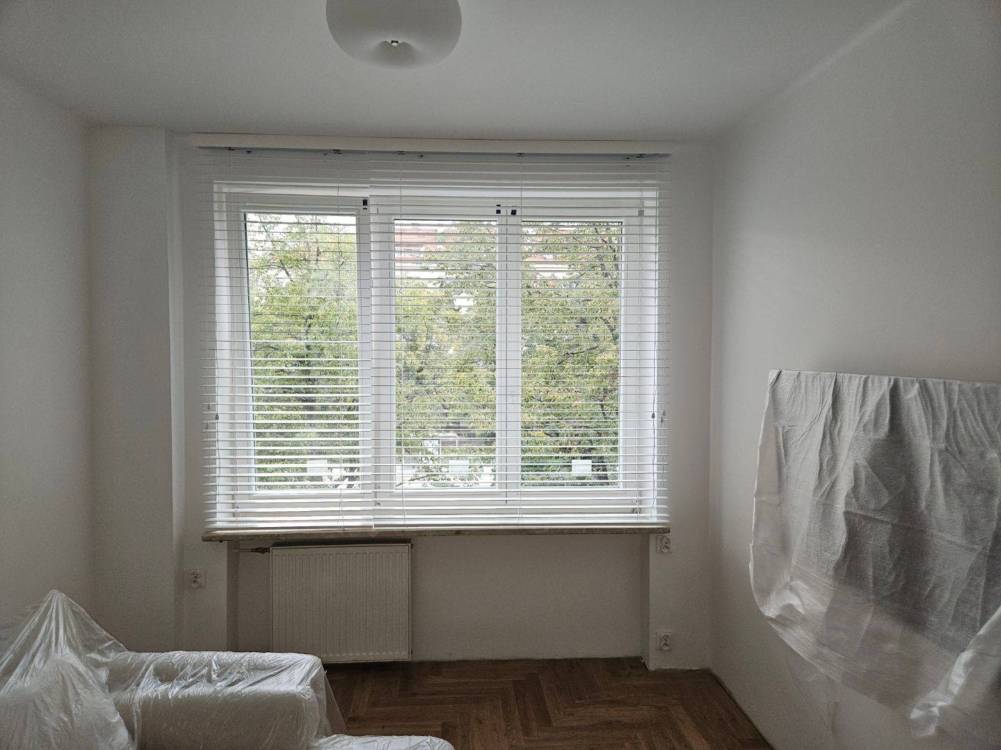 Wnętrze pokoju z oknem zasłoniętym białymi żaluzjami poziomymi, meble zabezpieczone folią malarską, widok na drzewa za oknem.