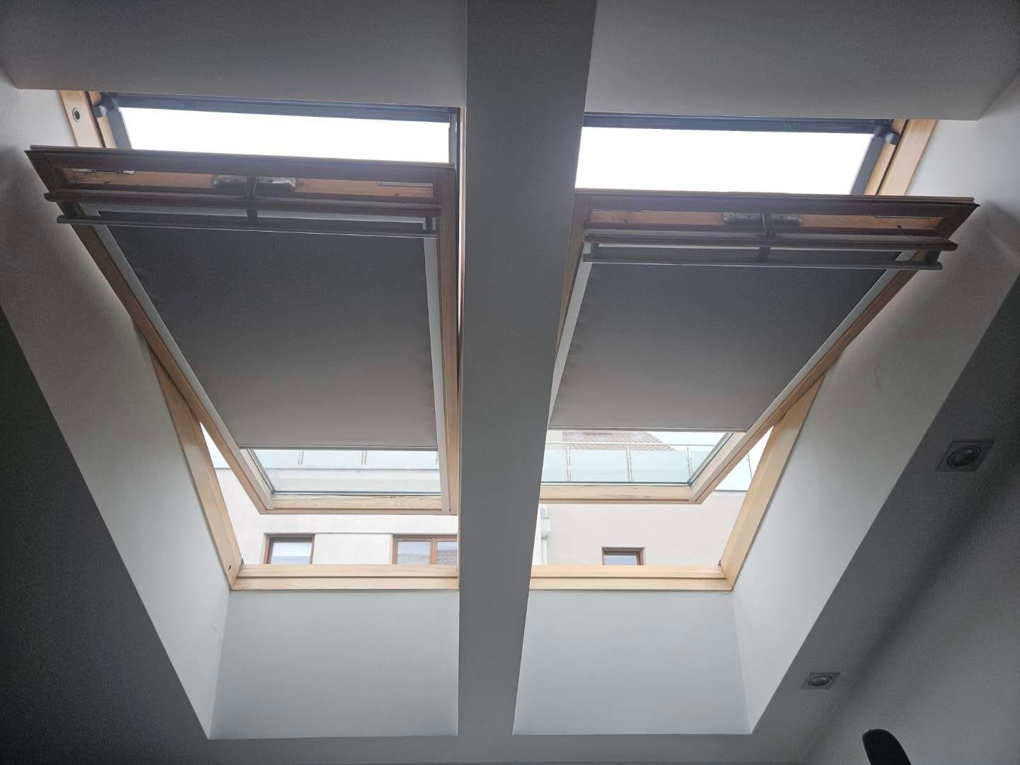 Dwa okna dachowe z drewnianą ramą i częściowo zasłoniętymi szarymi materiałowymi osłonami, widok na zewnątrz z fragmentami budynków.