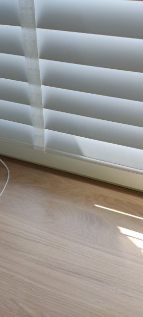 Fragment białych, poziomych osłon okiennych z widocznym mechanizmem sznurkowym, zamontowanych przy oknie, z fragmentem drewnianej podłogi w tle.