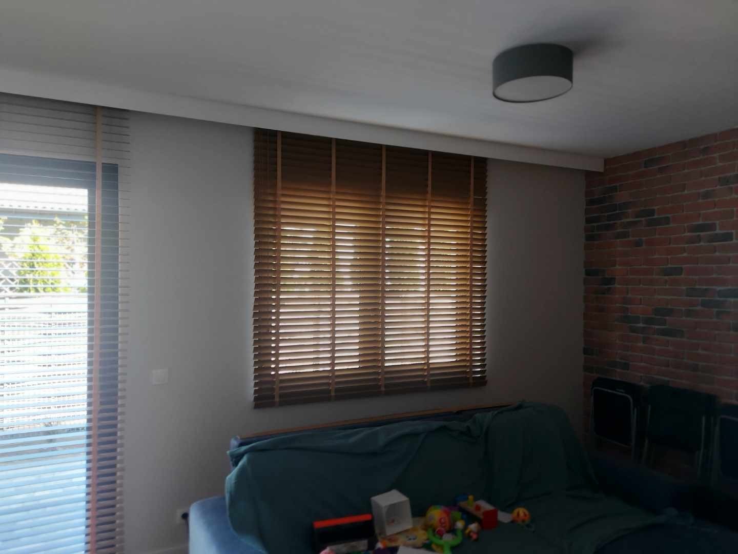 Brązowe poziome lamele okienne zamontowane w salonie z ceglaną ścianą, widoczne zabawki na sofie.
