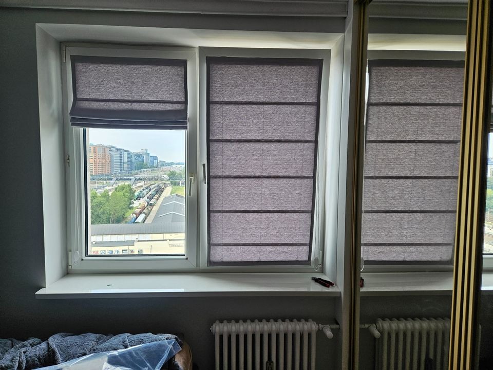 Dwa okna z szarymi tekstylnymi osłonami, jedno częściowo podniesione, widok na miasto z pociągami w tle, fragment grzejnika i szafy z lustrem w pomieszczeniu.