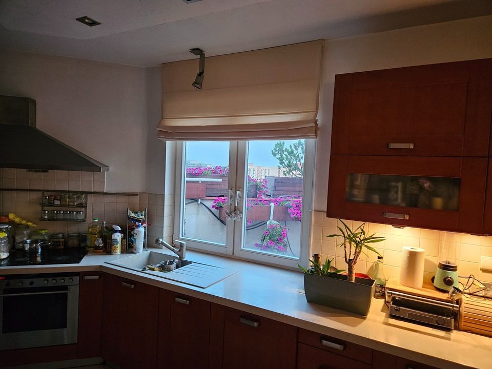 Kuchnia z drewnianymi szafkami i beżową roletą rzymską, za oknem balkon z różowymi kwiatami.