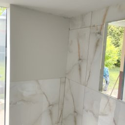 Jarosław Mikołajczyk - Ściany łazienki wyłożone białymi płytkami imitującymi marmur, z widocznym fragmentem umywalki i lustra, w którym odbija się postać pracownika na zewnątrz.