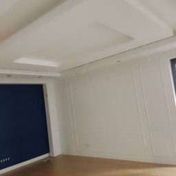 Jarosław Mikołajczyk - Eleganckie wykończenie wnętrza: białe ściany z dekoracyjnymi panelami, drewniana podłoga i sufit z kasetonami. Jedna ściana pomalowana na granatowo.