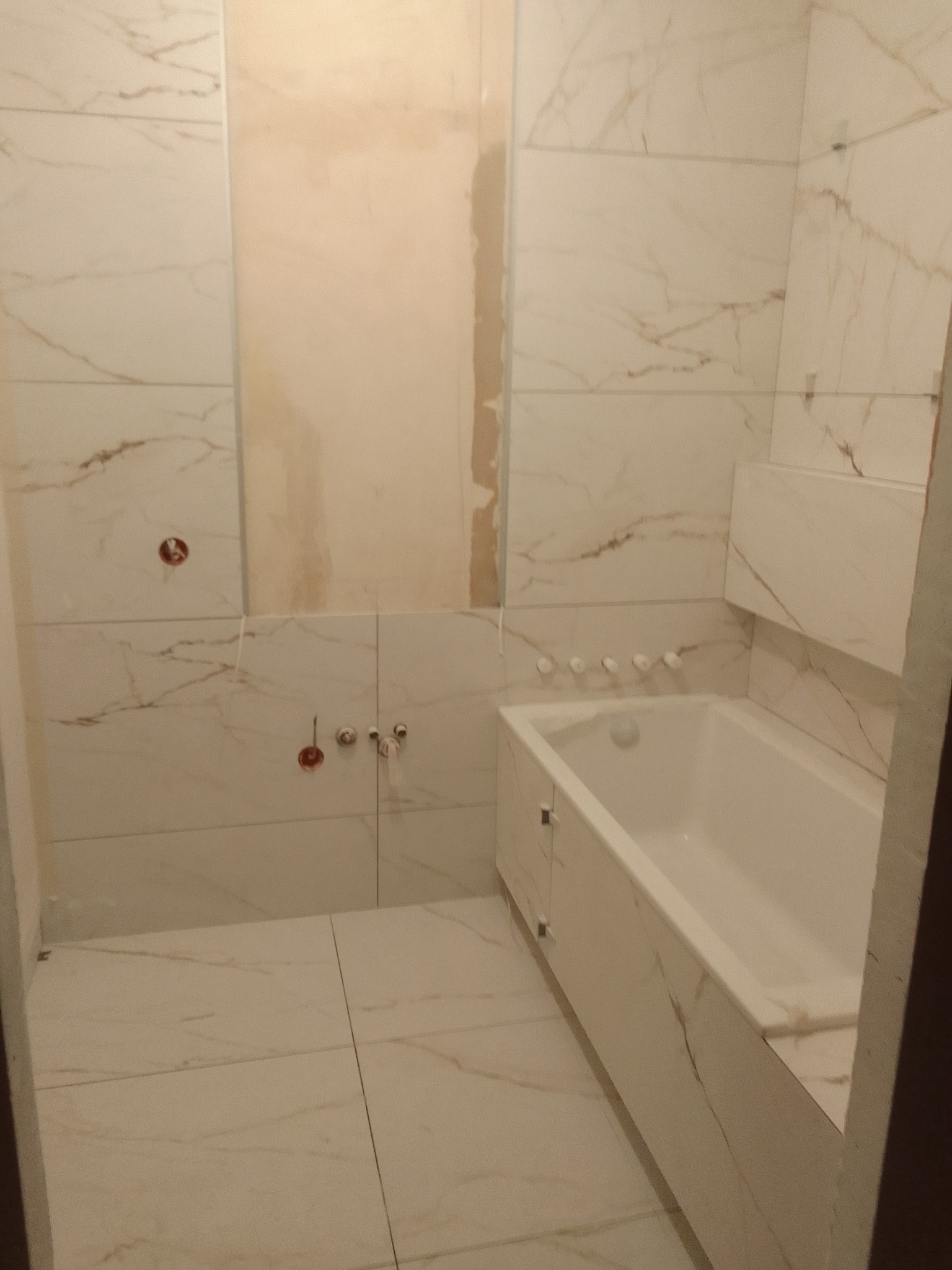 Wnętrze łazienki w trakcie prac wykończeniowych, widoczne płytki imitujące marmur na ścianach i podłodze, niezamaskowane przyłącza wodne oraz obudowana wanna z białym frontem.