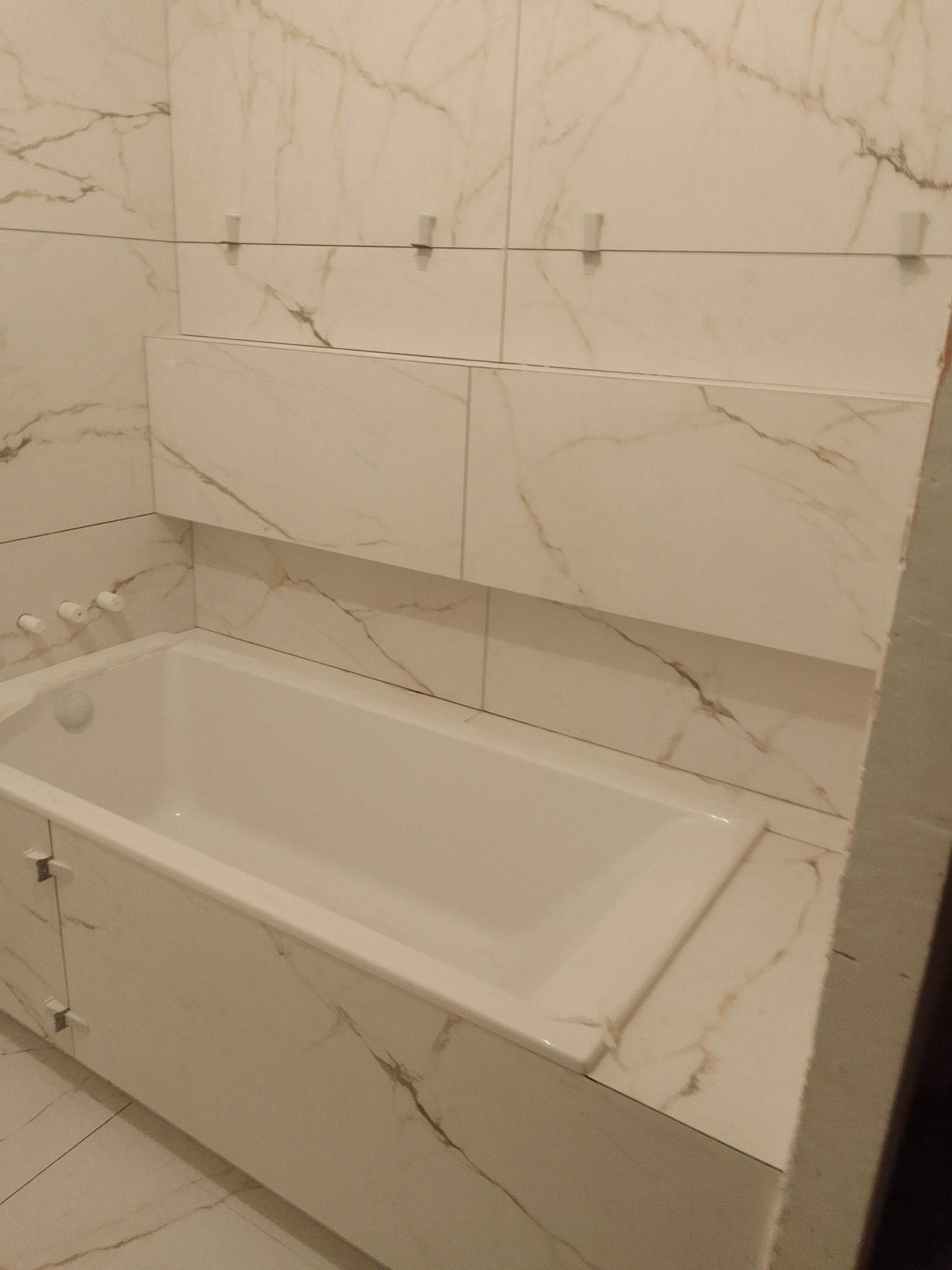 Wykończona łazienka z wanną obudowaną płytkami imitującymi marmur, widoczne białe płytki z brązowymi żyłami, listwy przypodłogowe oraz uchwyty do mocowania płytek.