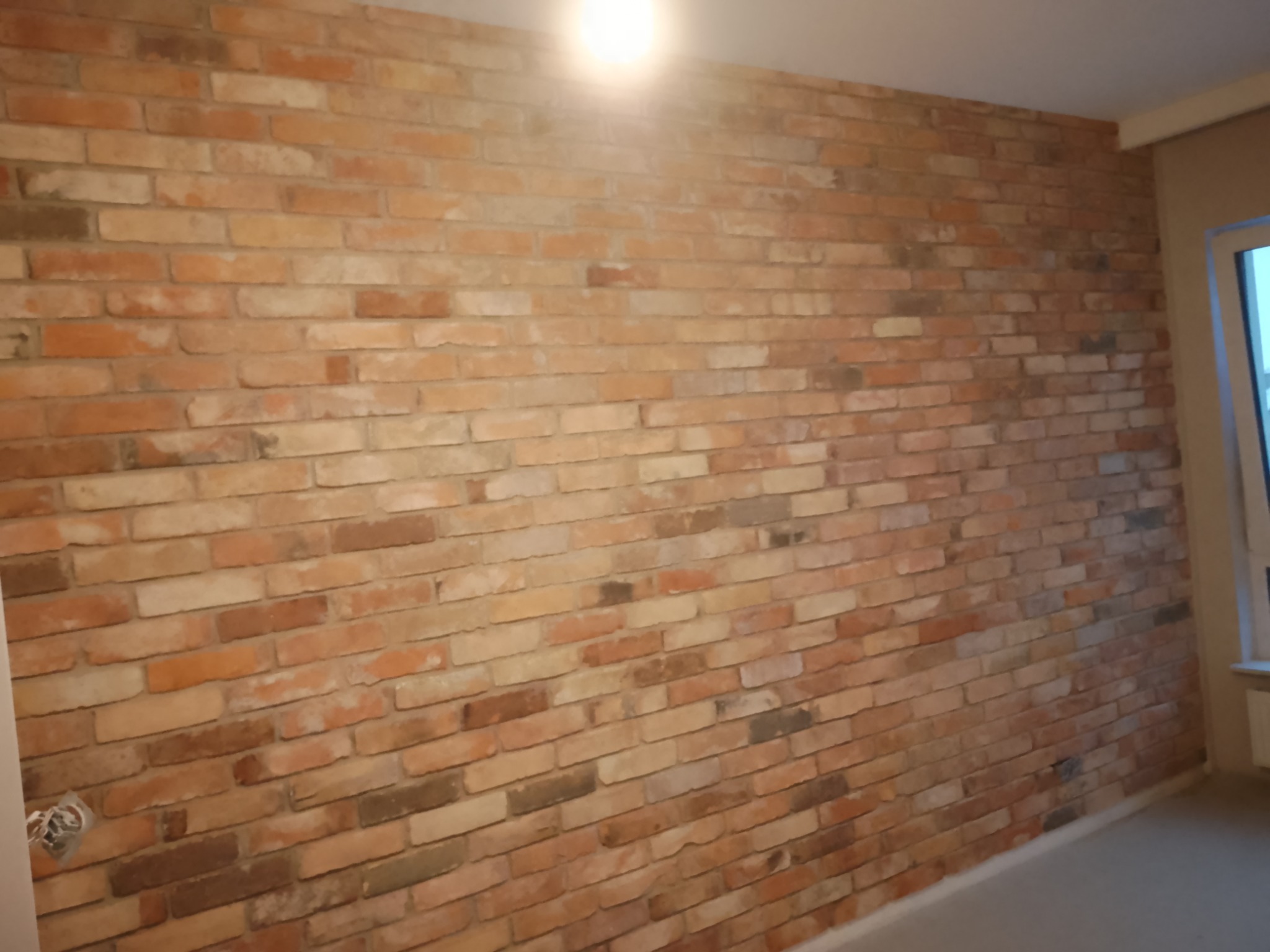 Ściana wyłożona cegłą dekoracyjną w odcieniach czerwieni i brązu, widoczna puszka elektryczna z przewodami, oświetlona punktowym światłem sufitowym.