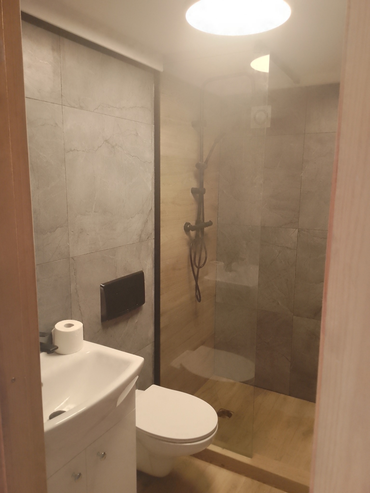 Nowoczesna łazienka z kabiną prysznicową typu walk-in, wykończona płytkami imitującymi marmur, z wiszącą toaletą i umywalką z szafką, oświetlona górnym światłem.