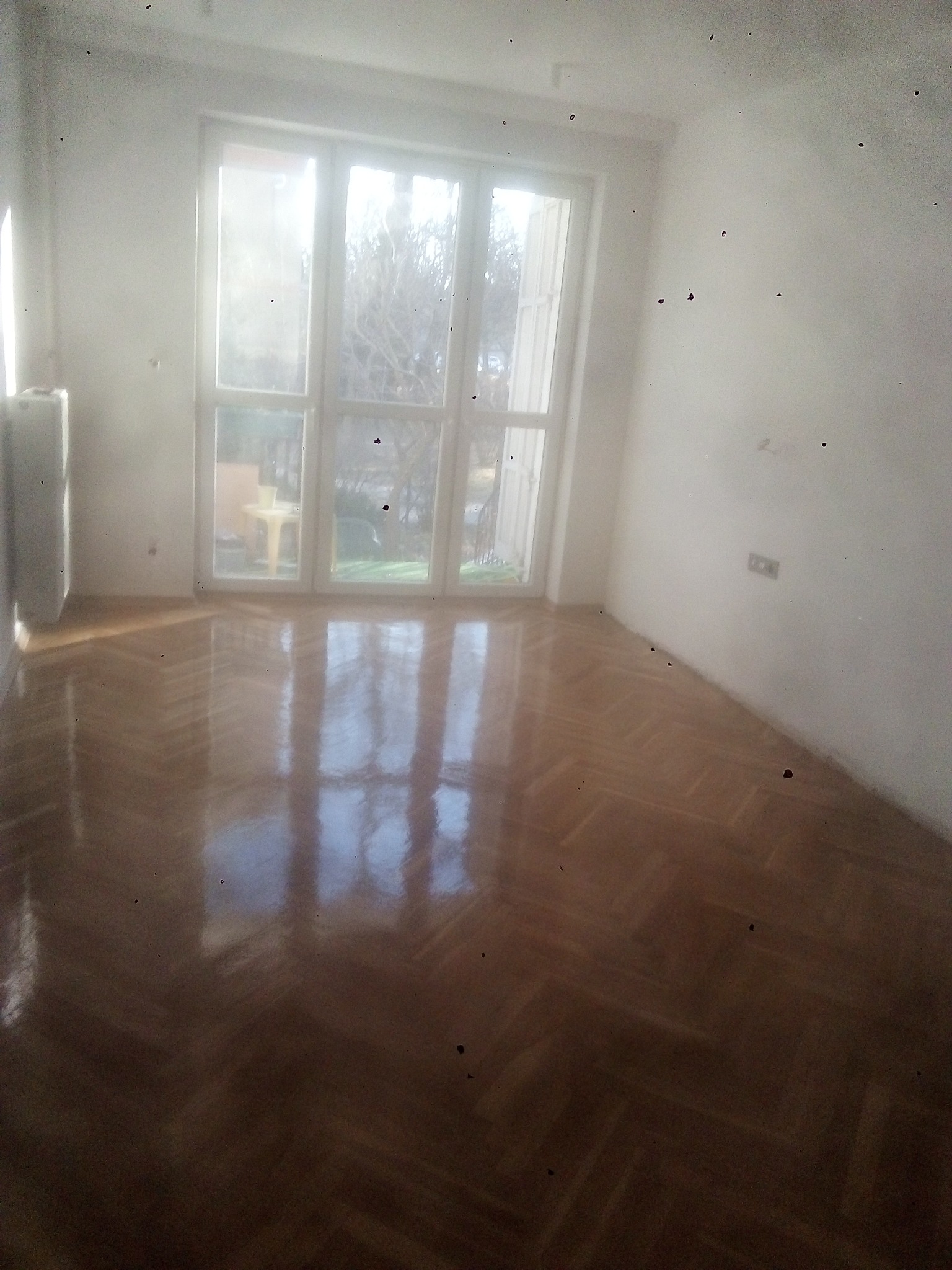 Pusty pokój z parkietem ułożonym w jodełkę, świeżo pomalowanymi na biało ścianami i dużym oknem balkonowym z widokiem na zewnątrz.