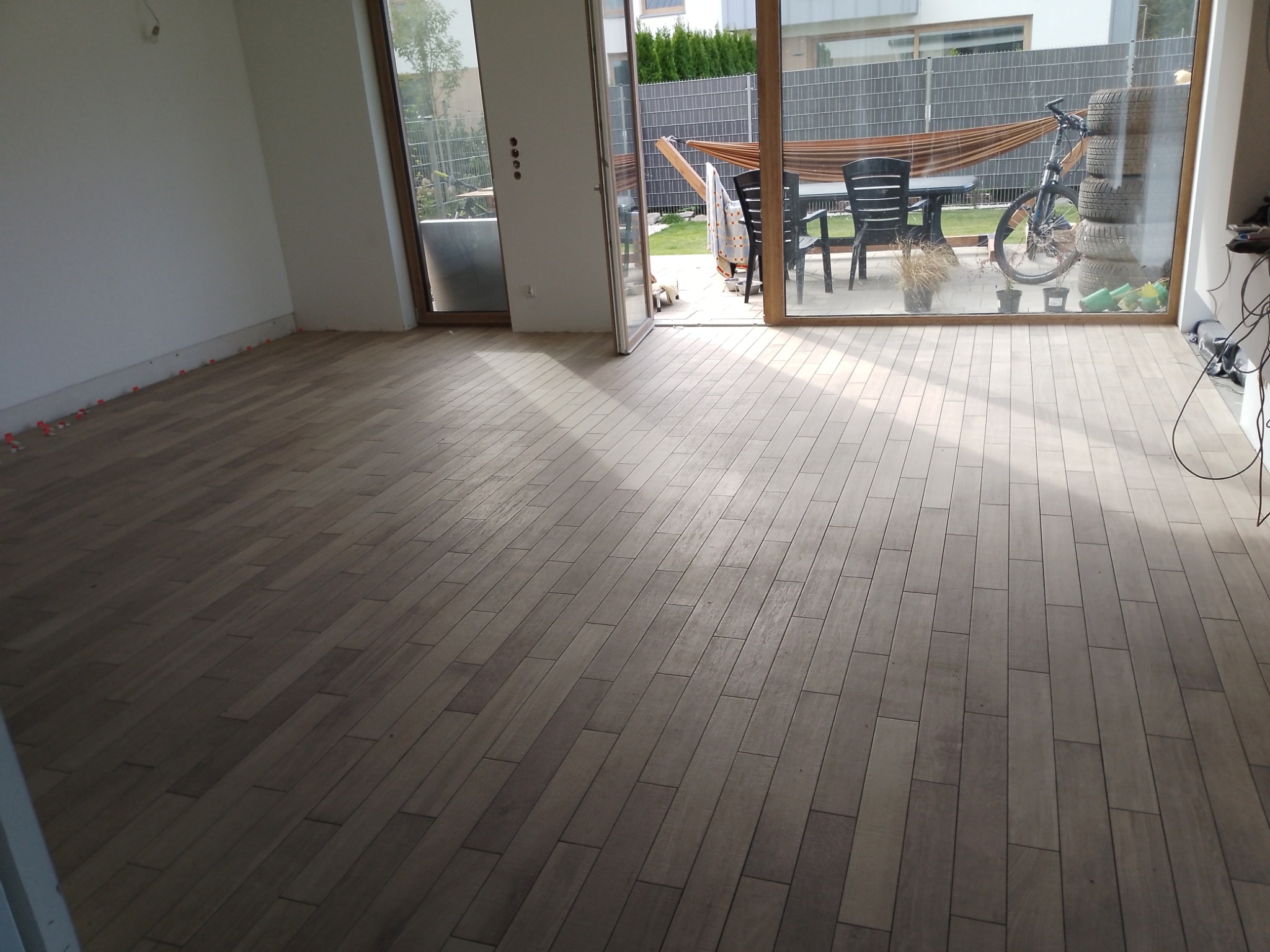 Ułożona podłoga z paneli imitujących drewno, widoczne plastikowe krzyżyki dystansowe przy ścianie, duże okno przesuwne z widokiem na taras z hamakiem i rowerem.